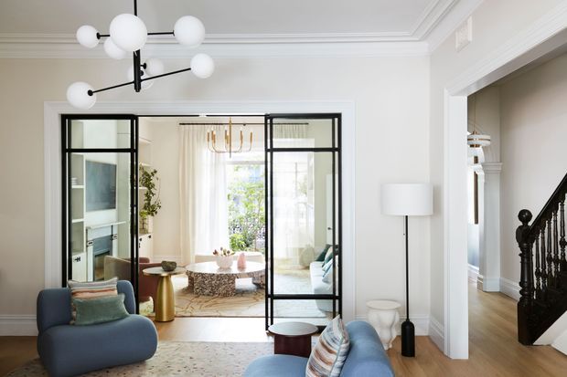 Bright, refined contemporary living room with a blue curved lounge chair and sofa, patterned rug, sculptural side tables, and a large black-and-white globe pendant. Black-framed glazed internal doors open toward a light-filled adjoining sitting area, while a traditional dark timber staircase and ornate white ceiling mouldings add classic architectural detail.