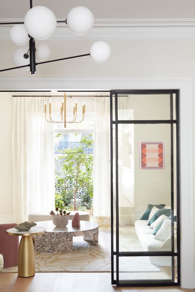 Bright, refined interior view through a black-framed glazed partition. The foreground features a sculptural white globe pendant, a brass side table, and a round marble coffee table with a substantial patterned stone base. Beyond the partition is a light-filled sitting area with a white sofa, teal cushions, sheer curtains, a brass chandelier, and framed artwork. Pale walls with decorative moulding, timber flooring, and a soft patterned rug create an elegant contemporary-classic atmosphere.