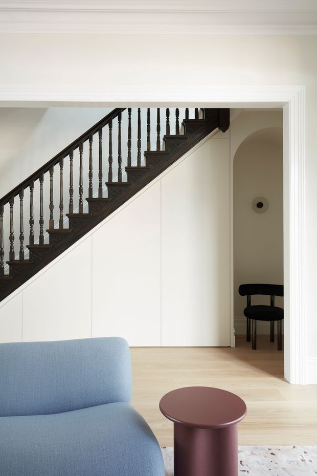 A bright, refined interior view of a living room with a pale blue curved sofa and small burgundy round side table in the foreground. A dark stained timber staircase with turned balusters runs diagonally across the rear wall, above flush white built-in cabinetry. A wide trimmed opening leads to a small adjoining alcove with an arched entry and black chair. Pale timber flooring, white walls, and layered white door casing create a clean contemporary setting with traditional stair detailing.