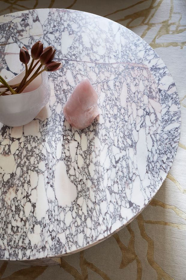 Close-up view of a round coffee table with a dramatic white marble top featuring dense grey and burgundy veining. A translucent white vessel with dark tulip stems and a pale pink decorative stone sit on the tabletop. The table is positioned over a light fabric rug with an abstract gold and taupe pattern.