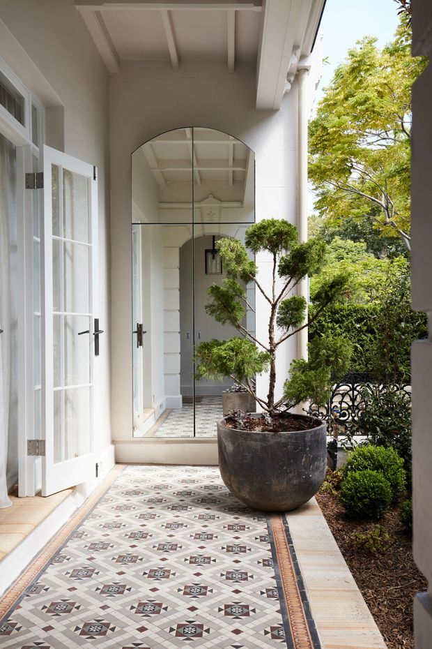 A bright covered entrance veranda or courtyard walkway with white-painted walls, exposed ceiling beams, tall French-style glazed doors, and a large arched mirror reflecting the corridor. The floor is finished with an intricate patterned tile mosaic, while a large potted topiary tree and manicured planting sit beside an ornate dark metal balustrade. The space has a refined heritage-inspired character with abundant natural light.