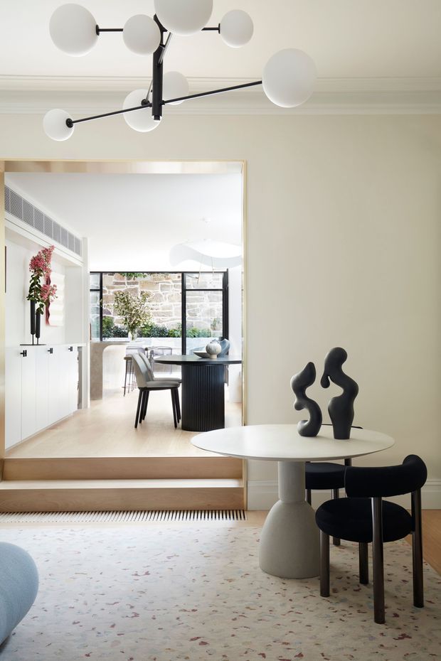 Refined contemporary interior showing a dining area in the foreground with a round white table, sculptural black chairs, and an abstract black ceramic sculpture. A large framed opening leads up several timber steps to a bright kitchen and dining space with pale cabinetry, stone walls, black-framed glazing, and indoor planting. The room features a modern globe chandelier, pale neutral finishes, decorative ceiling mouldings, and a soft patterned rug.