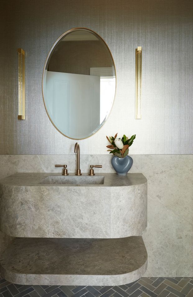A refined bathroom vanity with a curved, monolithic stone basin and floating lower shelf. The space features pale stone wall cladding, a textured neutral wall covering, brushed brass wall lights and tapware, an oval brass-framed mirror, and dark patterned floor tiles. A small floral arrangement adds a natural accent.