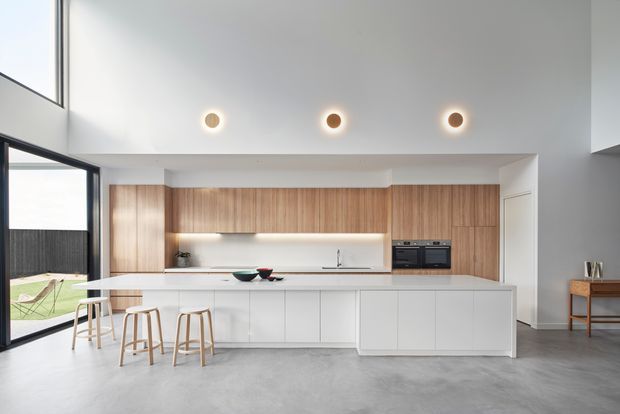 A bright, minimalist open-plan kitchen with a long white island and integrated cabinetry, warm vertical timber wall cabinets, a pale stone benchtop, and three timber stools. Full-height glazing and a glass door open to a small outdoor area, while recessed and under-cabinet lighting create a clean contemporary interior.