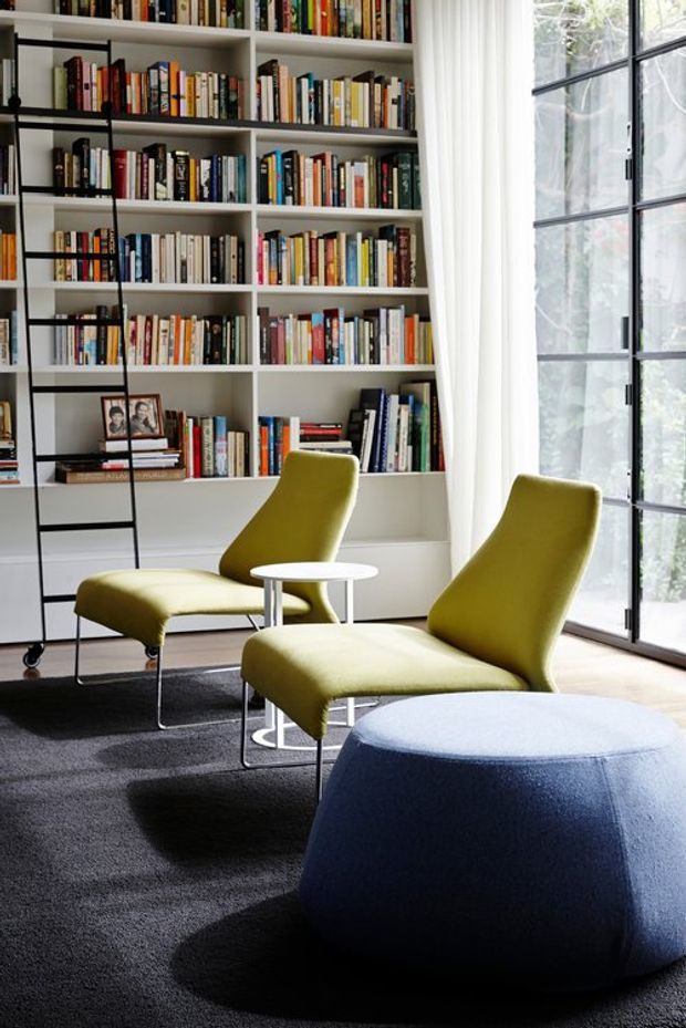 A bright, contemporary reading and living area with a large built-in white bookcase filled with books, a black rolling library ladder, two olive-green upholstered lounge chairs, a small round white side table, and a substantial blue upholstered ottoman. Floor-to-ceiling black-framed glazing with sheer curtains brings in abundant natural light, while dark carpet contrasts with the pale timber floor near the window.