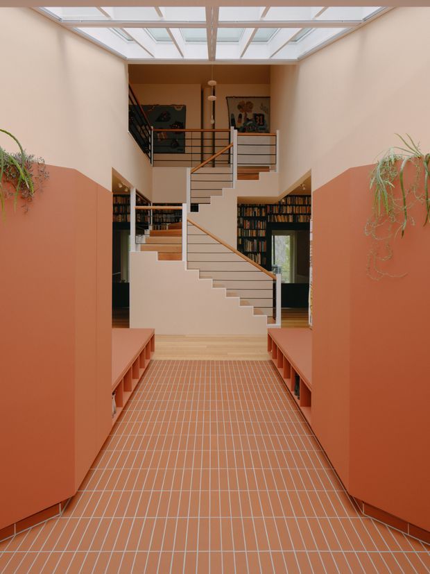 A dramatic interior stair hall with a central sculptural staircase leading to an upper landing. The space has terracotta-painted lower walls and built-in bench seating, pale peach upper walls, a terracotta grid-pattern tiled floor, dark built-in bookshelves, black and timber stair railings, and abundant overhead skylights. Hanging plants and artwork add character, while the strong axial composition emphasizes the double-height volume.