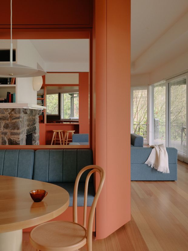 Warm, carefully composed open-plan interior showing a dining area in the foreground, a lounge beyond, and a kitchen/bar zone in the background. The space features salmon-red painted structural or partition walls, pale timber flooring, a rounded timber dining table and bentwood chair, a blue upholstered banquette and sofa, a stone-clad fireplace or kitchen feature, extensive windows with horizontal blinds, and a sculptural white pendant light. The interior has a refined mid-century-inspired character with strong colour blocking and curved furniture forms.