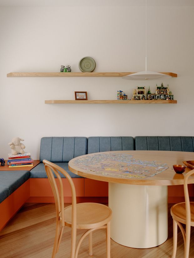 A warm, compact dining nook with a built-in L-shaped banquette upholstered in blue-grey fabric, orange cabinetry below, and a round pale wood dining table with a cylindrical light-toned pedestal. Bentwood dining chairs surround the table. Two long floating timber shelves display small decorative objects and collectibles, while a slim white pendant light hangs above. The room has pale walls, timber flooring, and a playful yet refined mid-century-inspired interior.