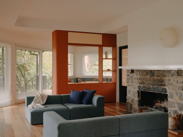 A warm, retro-inspired living room with a large sectional sofa facing a substantial natural-stone fireplace. The room features timber flooring, expansive window and glazed-door openings, and a striking built-in reddish-brown timber joinery wall incorporating shelving, cabinetry, and a framed view into an adjacent dining area. Soft neutral walls, a sculptural wall light, and dark blue cushions complete the relaxed interior.