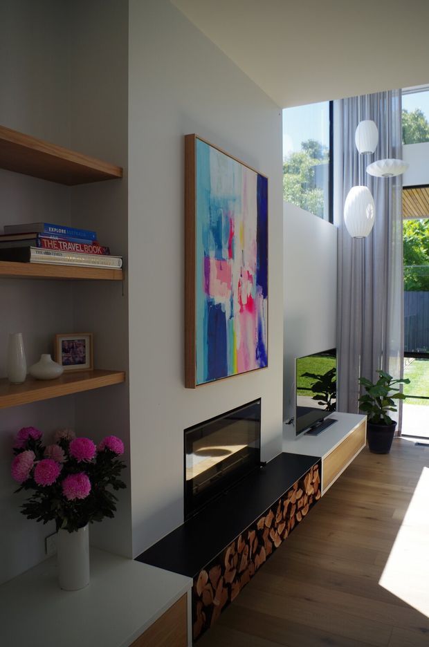 A tall, light-filled contemporary living room with a double-height glazed opening, sheer curtains, timber flooring, and a built-in media and fireplace wall. The feature wall includes open timber display shelving, a colourful abstract artwork, a black fireplace surround, stacked firewood storage, and a low timber TV console. Sculptural pendant lights and indoor plants add decorative detail.