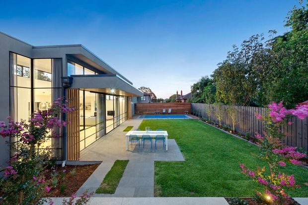 A contemporary single-storey home opens onto a landscaped backyard with extensive floor-to-ceiling glazing, a covered outdoor dining terrace, paved paths, lawn, timber fencing and a compact rectangular swimming pool. The flat-roofed building has dark grey exterior finishes and warm interior lighting, with flowering planting framing the garden.