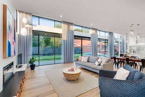 Bright contemporary open-plan living and dining area with floor-to-ceiling black-framed glazing, sheer curtains, pale timber flooring, a neutral sectional sofa, patterned armchairs, and a sculptural round coffee table. A fireplace and wall-mounted artwork sit to the left, while the dining area and kitchen are visible toward the rear. The room opens visually to a landscaped garden through extensive glazing.