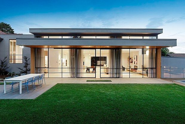 Contemporary two-storey residential home with a strong horizontal profile, broad flat roof overhangs, extensive floor-to-ceiling glazing, and warm timber detailing. The rear facade opens onto a manicured lawn and paved outdoor dining area, with bright, minimalist interiors visible through the glass.