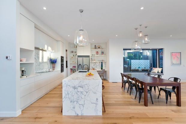 Bright open-plan kitchen and dining area with white built-in cabinetry, a large white veined stone island, timber flooring, pendant lighting, and a dark timber dining table. Glazed doors at the rear open toward an outdoor area, while integrated shelving and framed artwork add detail to the space.