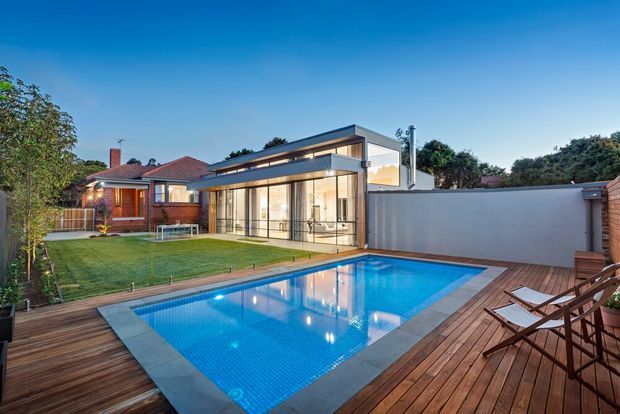 Exterior view of a contemporary glass-walled pool pavilion or extension beside a landscaped backyard swimming pool. The scene includes a timber pool deck, blue tiled pool, white rendered boundary wall, and an older red-brick tiled-roof house in the background at dusk.