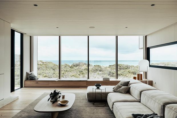 A bright, refined living room with a large cream sectional sofa, built-in timber window seat, low organic-shaped coffee table, and expansive floor-to-ceiling glazing overlooking coastal vegetation and the ocean. The room has a pale timber floor, white vertically panelled walls, a subtly ribbed white ceiling, and slim dark window frames.