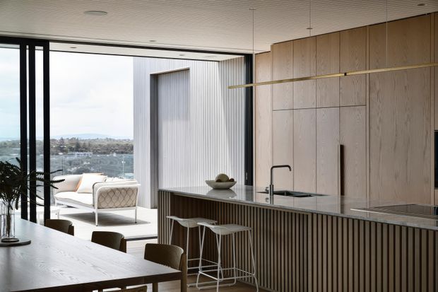 Contemporary open-plan kitchen and dining area with a large timber island, integrated light-wood cabinetry, dark vertical slatted detailing, and a minimalist dining table. Full-height black-framed glazing opens to an outdoor terrace with a view beyond. The space has a restrained, refined palette and abundant natural light.