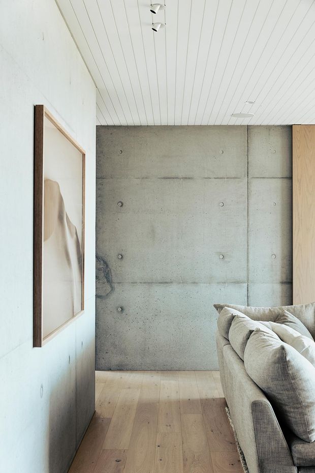 A refined contemporary living room corner with a large concrete feature wall, pale timber flooring, white painted vertical-panel ceiling, and a neutral upholstered sofa with cushions. A framed artwork hangs on the white wall, while warm timber joinery is visible at the right edge. The space has a restrained material palette and exposed concrete detailing.