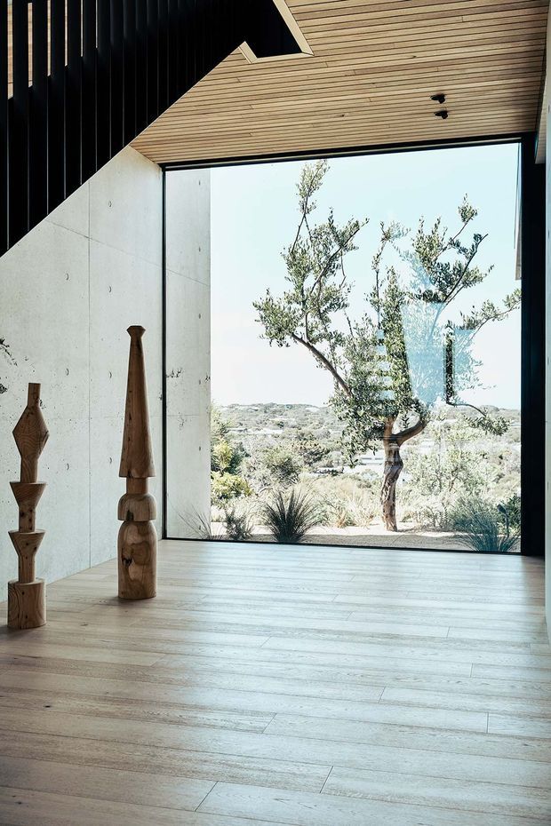 Minimalist interior with pale timber flooring, a large black-framed picture window, white concrete walls, and a slatted timber ceiling. Sculptural wooden objects stand near the window, which overlooks a dry landscaped setting with native-style vegetation. A dark vertical slatted element and angular ceiling opening add architectural contrast.