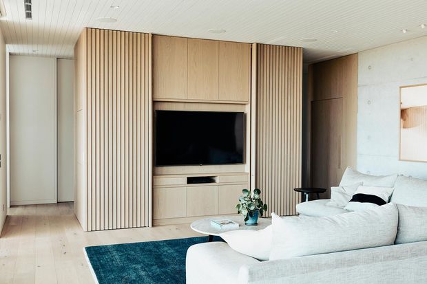 Bright contemporary living room featuring a large pale upholstered sectional sofa, dark teal area rug, light timber built-in media cabinetry, vertical timber slat wall panels, and a wall-mounted television. The space has pale timber flooring, a white slatted ceiling, and a raw light concrete feature wall with a doorway opening to another room.