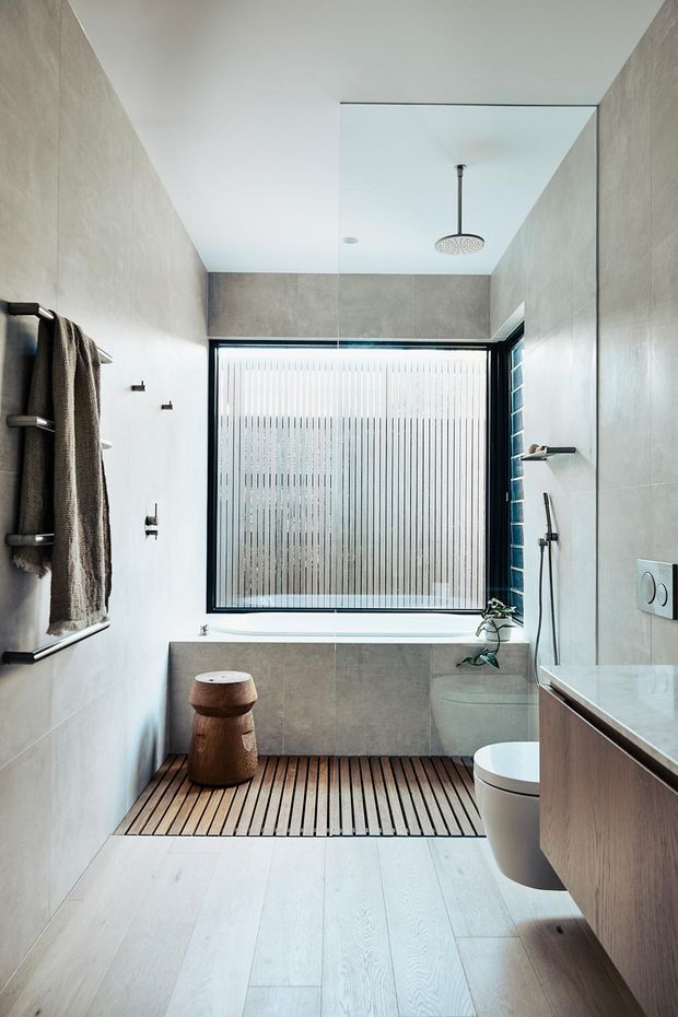 Bright contemporary bathroom with large-format pale grey wall tiles, a freestanding rainfall shower, frameless glass shower screen, wall-mounted vanity, concealed toilet, bathtub beneath a textured privacy window, timber slatted bath mat/decking, and dark timber and black metal accents. A small wooden stool and towel rail add warmth to the minimalist space.