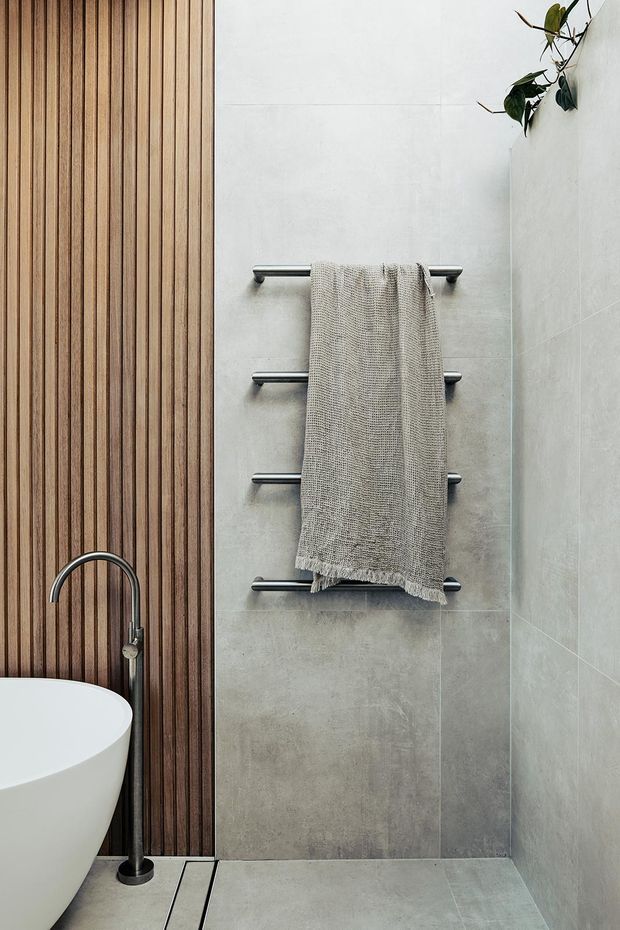 A contemporary bathroom with a freestanding white bathtub partially visible at the lower left, a dark brushed-metal floor-mounted bath tap, pale large-format tiled walls and floor, and a vertical timber slat feature wall. A minimalist chrome towel rail holds a neutral textured towel.