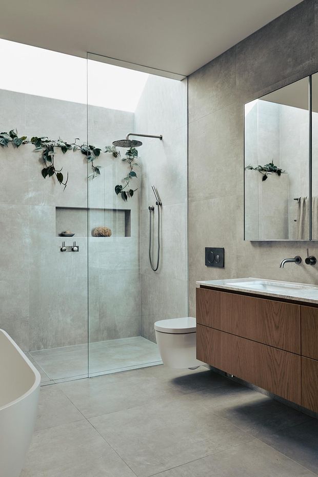 Contemporary bathroom with a frameless glass walk-in shower, large grey concrete-look tiles, wall-mounted toilet, freestanding bathtub, and a floating dark timber vanity with an integrated white basin. A large mirror, recessed shower niche, black fixtures, and trailing greenery complete the refined, natural-toned interior.