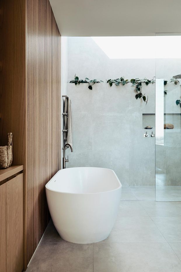 A refined modern bathroom featuring a freestanding white bathtub, large pale stone-look floor and wall tiles, a frameless glass shower enclosure, and a warm vertical timber slat wall with built-in cabinetry. A narrow ledge with trailing greenery adds a natural accent, while a skylight or clerestory opening provides soft daylight.