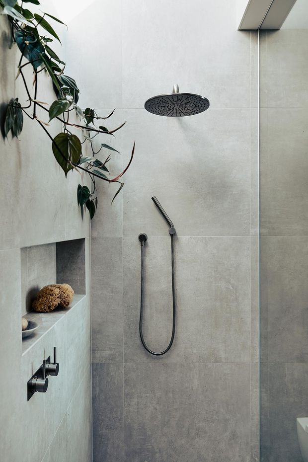 A contemporary walk-in shower with large-format light grey stone-look wall tiles, a black rain showerhead and handheld shower, recessed wall niche, and black fixtures. A trailing indoor plant adds a natural accent, while a partial toilet is visible at the lower right.