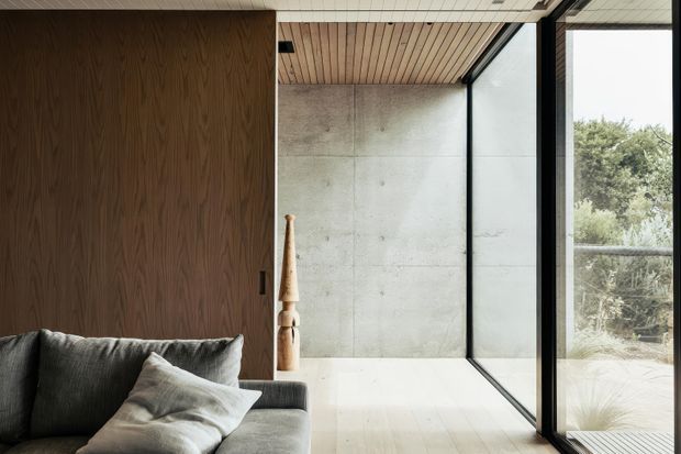 A refined modern living room corner with a dark grey upholstered sofa and cushions in the foreground, a full-height walnut-toned timber wall panel, pale concrete feature wall, light timber slatted ceiling, and expansive black-framed glazing opening toward landscaped greenery. The restrained material palette and strong indoor-outdoor connection create a calm, contemporary atmosphere.