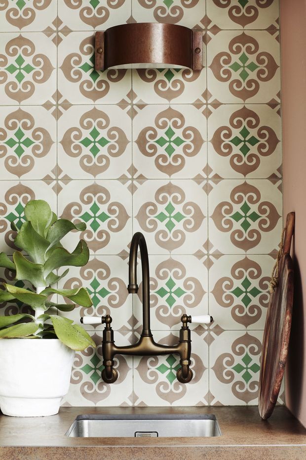 Close-up of a compact kitchen sink area featuring a patterned ceramic tile splashback in cream, taupe and green, a dark antique-brass wall-mounted gooseneck tap with white lever handles, a stainless-steel undermount sink, stone-look benchtop, wall sconce, potted plant and leaning timber board.