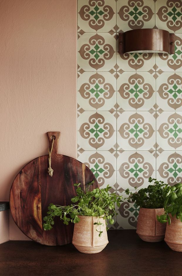 A close-up styled interior vignette featuring a patterned ceramic tile wall in cream, taupe, and green, paired with a muted terracotta plaster wall. A dark wooden cutting board leans against the wall beneath a small curved timber wall-mounted shelf or bracket, while potted leafy herbs and plants sit on a dark countertop or ledge. The composition has a warm, earthy, Mediterranean-inspired feel.