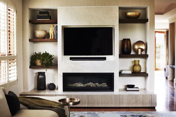 A refined living room centered on a built-in entertainment wall with a large flat-screen television above a linear fireplace. Open dark timber display shelves flank the pale stone or plaster feature surround, styled with ceramics, books, plants, and sculptural objects. A low timber media cabinet, neutral upholstered seating, plantation shutters, and polished timber flooring create a warm, sophisticated interior.