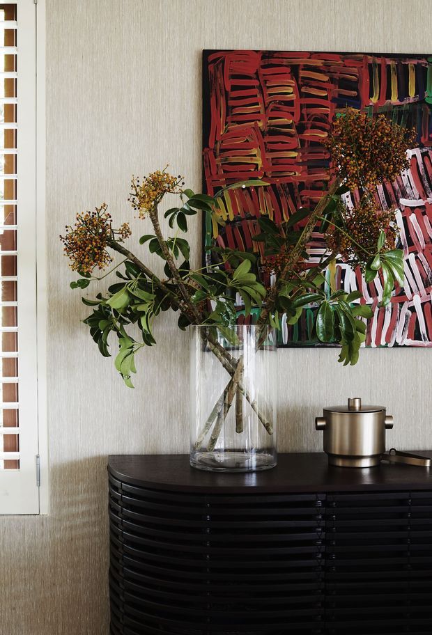 A styled interior vignette featuring a black curved slatted sideboard against a textured beige wall. A large clear glass vase holds leafy flowering branches, positioned in front of a bold abstract artwork in black, red, orange, pink and green. A metallic cylindrical decorative vessel sits on the sideboard, while a white-framed glazed door or screen is visible at the left edge.