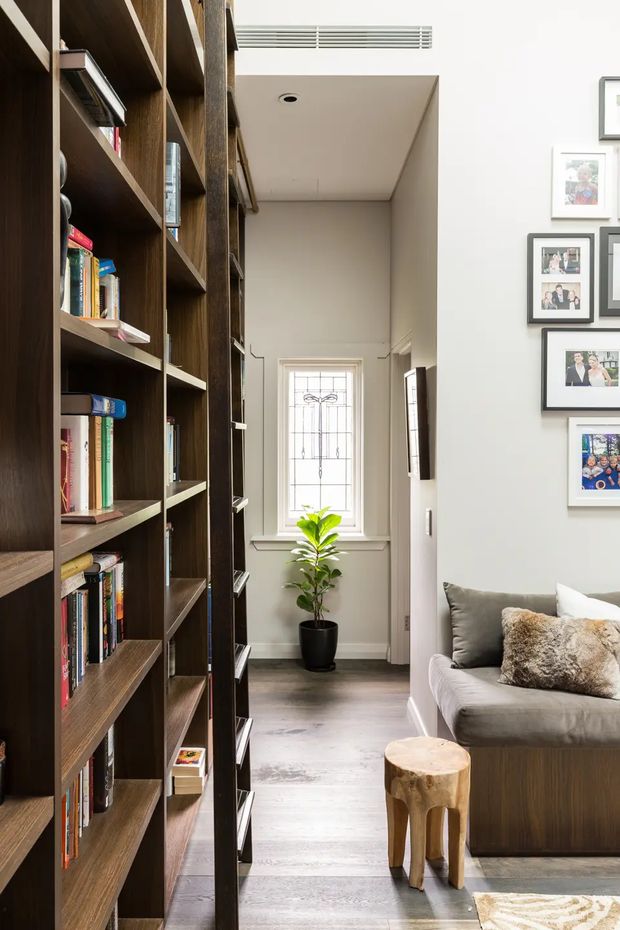 A bright, narrow living room or reading area with a tall, dark timber built-in bookshelf forming a strong architectural partition on the left. A grey sofa with textured cushions sits on the right beside a small natural timber stool, while a decorative leadlight-style window and potted plant are visible at the end of the space. Family photographs are arranged on the pale wall, and light timber flooring, recessed lighting, and a linear ceiling vent complete the room.
