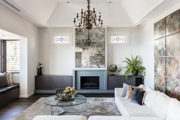 A refined living room with a white vaulted coffered ceiling, dark timber flooring, a substantial white sectional sofa, glass coffee table, dark built-in cabinetry, and a central contemporary fireplace surround. A decorative chandelier hangs over the seating area, while mirrored or metallic feature panels and framed wall artwork add visual interest. Decorative leaded-glass windows and classical detailing are balanced with contemporary furnishings and finishes.