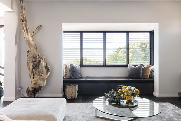 Bright contemporary living room with a built-in black window seat beneath expansive horizontal windows, a pale upholstered chaise, round glass coffee table, textured rug, neutral grey walls, and a dramatic sculptural piece of natural timber. The room has strong daylight, clean lines, and restrained monochromatic styling with organic accents.