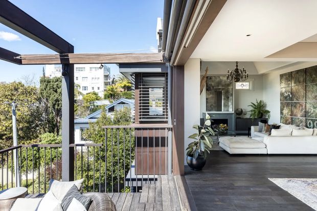 A bright contemporary indoor-outdoor living space opening onto a timber deck balcony. The room features dark timber flooring, a white sectional sofa, a black fireplace or media wall, decorative chandelier, indoor plants, and large artwork. Slim dark-framed glazing and structural posts connect the living room to the covered deck, which has timber decking and metal balustrades. The elevated urban-suburban outlook is visible beyond the railing.
