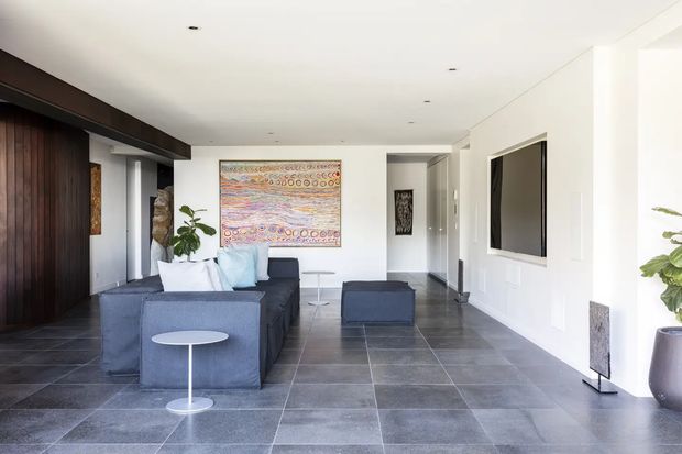 Bright contemporary living room with a low, streamlined charcoal sectional sofa and ottoman arranged around small white side tables. The room features large-format dark grey tiled flooring, white walls and ceiling with recessed downlights, dark timber built-in cabinetry or wall panelling on the left, and a wall-mounted flat-screen television on the right. A colourful abstract artwork provides a focal point, while indoor plants and minimal furnishings add softness. An open hallway and entry area are visible toward the rear.