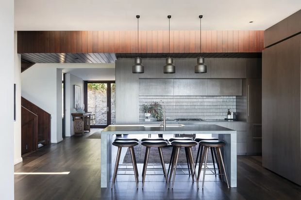 Contemporary open-plan kitchen viewed from the living area, featuring a large stone island with four bar stools, dark timber flooring, grey fixed cabinetry, a horizontally tiled splashback, and a warm timber-lined bulkhead with pendant lighting. A stair and entry area are visible beyond the kitchen.