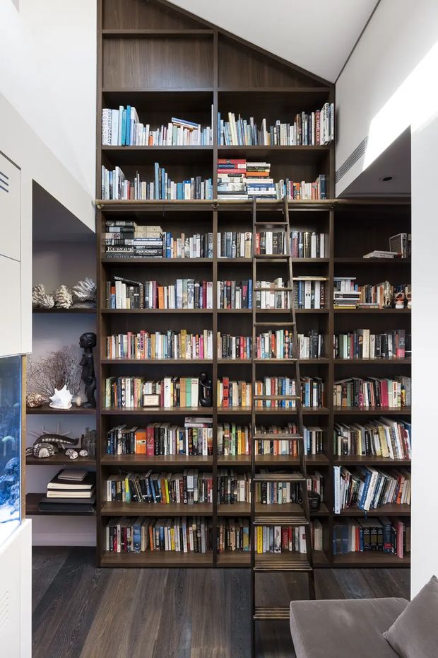 Tall built-in dark timber library shelving fills the wall from floor to ceiling, with numerous books arranged across open shelves and an integrated rolling library ladder. The shelving follows a sloped ceiling line at the top and includes a small display niche at the side. Dark timber flooring, white walls, and part of a neutral upholstered sofa are visible. The space is a refined, book-filled residential interior with strong custom joinery.