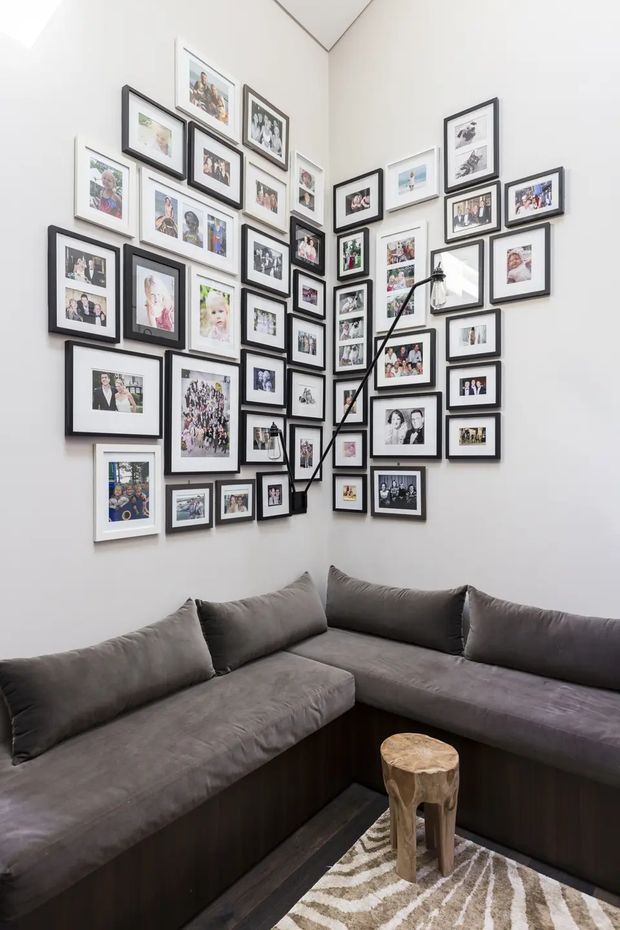 A compact, bright living room corner with a large dark-grey L-shaped sectional sofa built into the space. The white walls are densely arranged with a gallery of family photographs in mixed black and white frames, and a slim black floor lamp reaches into the corner. A small rustic wooden stool sits on a patterned neutral rug in front of the sofa. The room has a cozy, personal, contemporary apartment feel.