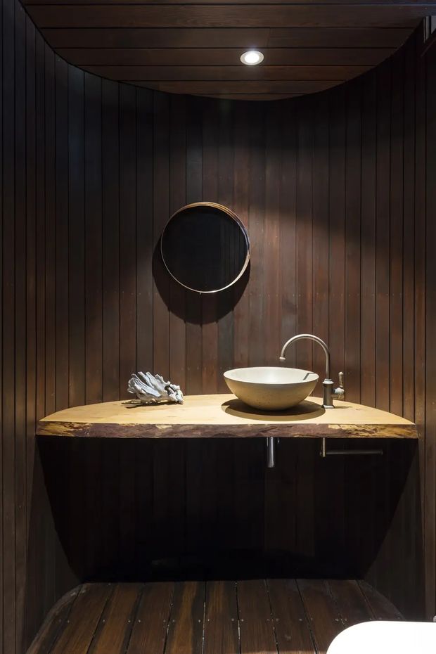 A compact, dark wood-lined bathroom with vertical timber wall panelling, a curved timber ceiling, and matching timber floorboards. A floating dark vanity supports a thick natural-edge light wood benchtop with a round vessel basin and brushed metal tapware. A round black-framed mirror and a recessed ceiling spotlight complete the moody, intimate setting.