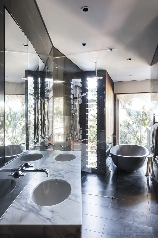 A luxurious contemporary bathroom with a double vanity, white veined stone benchtop, twin undermount basins, large mirrored wall panels, dark tiled flooring, a freestanding black bathtub, and a separate glass-enclosed shower. Floor-to-ceiling glazing brings in abundant natural light and views of palm foliage.