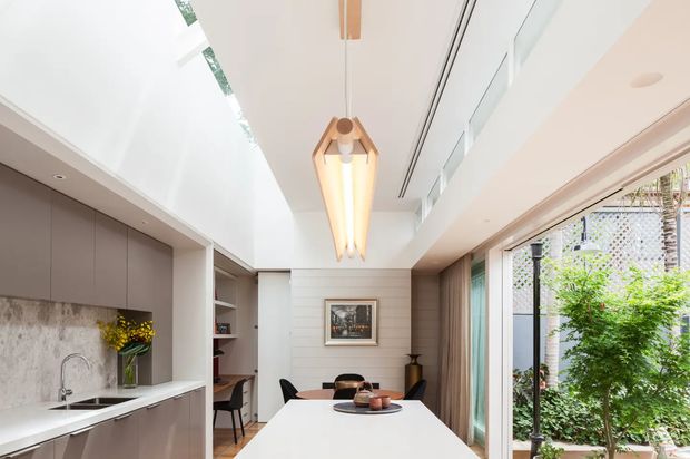 Bright contemporary open-plan kitchen and dining area with a long white island, integrated sink, taupe cabinetry, marble-look splashback, timber wall panelling and shelving. A dining table sits beyond the island beneath a sculptural pendant light, while full-height glazing opens toward a leafy courtyard or garden. The space has a clean, refined palette and strong natural light.