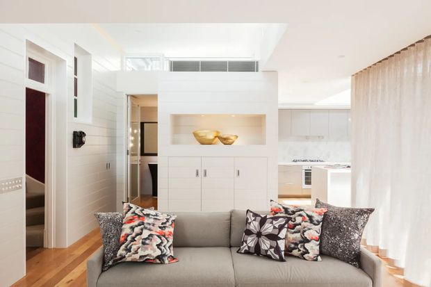 Bright contemporary open-plan living room viewed from behind a grey sofa, with white built-in cabinetry and recessed display niche, timber flooring, sheer curtains, and a partially visible white kitchen beyond. The room has abundant daylight and a clean, refined fit-out with patterned cushions providing small colour accents.