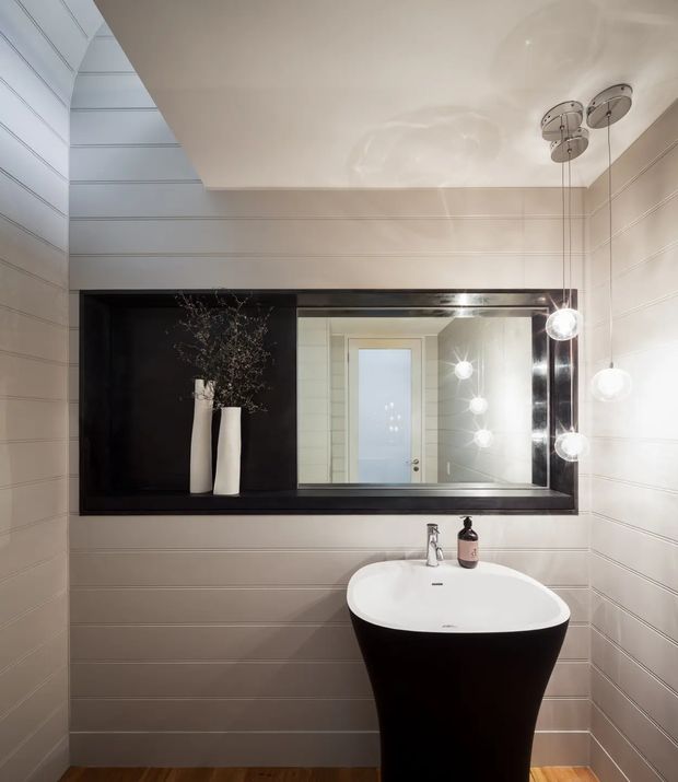 A compact, refined bathroom with horizontally panelled light walls, a dramatic black-framed recessed mirror and shelf, and a freestanding black pedestal basin with white top. Globe pendant lights hang beside the mirror, while warm timber flooring adds contrast.