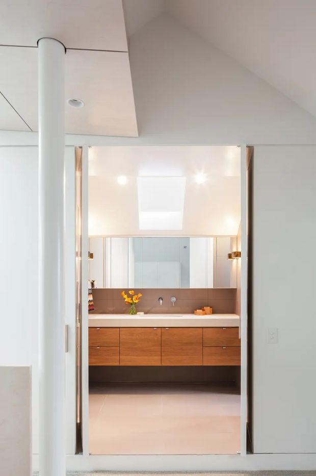 View through a doorway into a bright, contemporary bathroom with a floating timber vanity, white benchtop, broad mirror, warm brown backsplash, wall-mounted taps and integrated lighting. The foreground shows a white structural column and clean pale walls and ceiling.