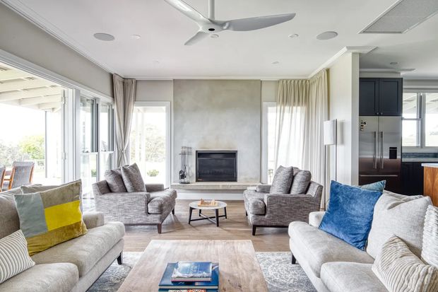 Bright, spacious contemporary living room with multiple upholstered sofas and armchairs arranged around a low coffee table and a built-in fireplace. Floor-to-ceiling glazed doors and windows open toward an outdoor area, with sheer curtains, a ceiling fan, pale timber flooring, and a neutral palette accented by blue and yellow cushions. A kitchen is partially visible through the opening at right.