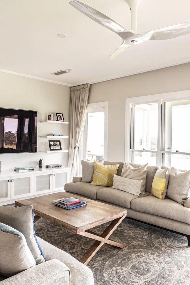 Bright, neutral-toned living room with a large sectional sofa, patterned area rug, rustic timber coffee table, built-in white media cabinetry and open shelving, framed photographs, and expansive windows with full-length curtains. A contemporary white ceiling fan and recessed lighting sit above the room, while yellow and blue cushions add restrained colour accents.